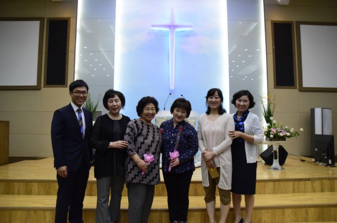 빛나는 십자가가 있는 강단 앞에서 꽃을 든 여섯 사람이 함께 선 단체 사진
