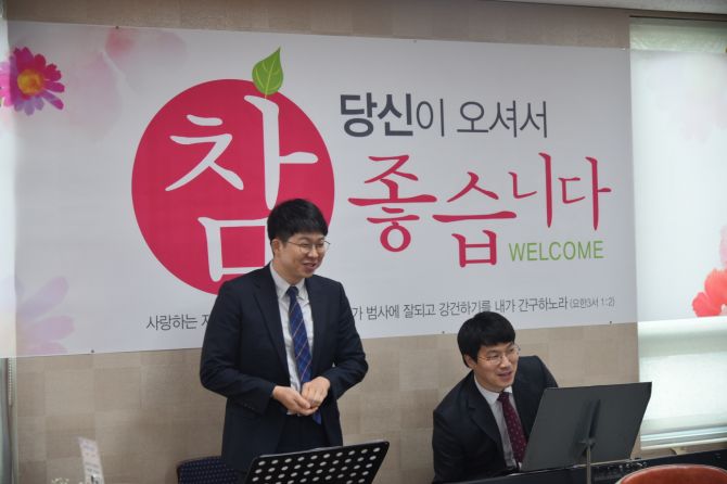 '당신이 오셔서 참 좋습니다' 환영 현수막 앞에 선 사람과 건반 앞에 앉은 사람