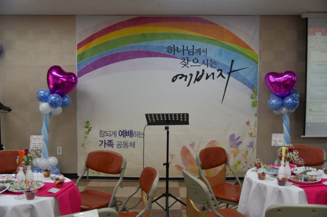 무지개 그림 현수막과 하트 풍선 장식 앞에 둥근 식탁과 의자가 놓인 행사장