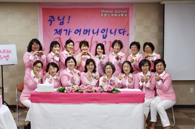 분홍 옷을 맞춰 입은 여러 사람이 꽃 장식 탁자 뒤에서 손으로 하트를 만들며 찍은 단체 사진